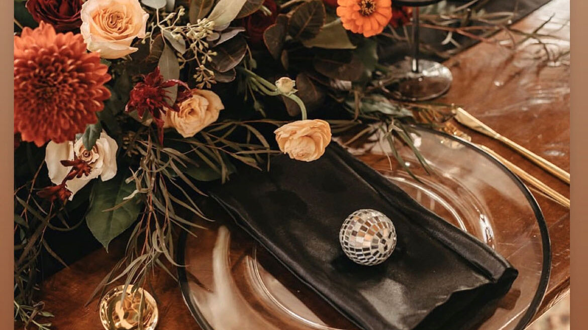 Fun and funky wedding table setting featuring bright seasonal fall colors and flowers, rich natural wood table, clear glass plates, and black flatware with disco ball charm