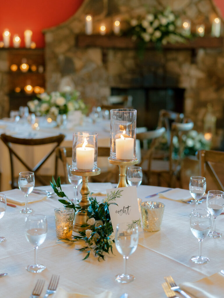 wedding centerpiece with pillar candles on gold candlesticks with hurricane glass, coordinating votives, and lush greens with tucked spray roses