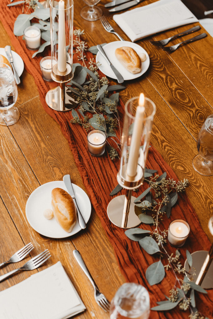 wedding centerpiece on a rustic wooden table with rusty orange cotton table runner, taper candles in tall gold candlesticks and hurricane glass, votives, and scattered eucalyptus greens