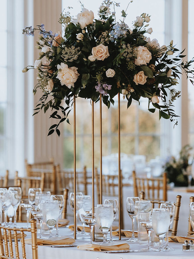 Tall wedding centerpiece set on a harlow stand featuring lush greens, ivory roses and peonies, and blue delphinium. 
