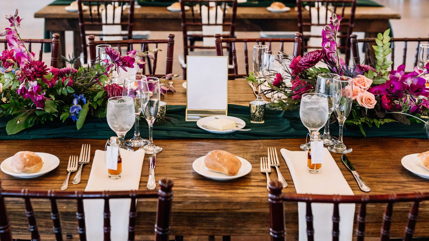 wedding banquet table with runner-style centerpiece including rich green cloth draping, cheerful and bright flowers in shades of pink, and table setting with white linens and wedding favors at each setting 